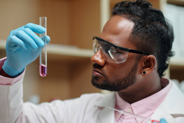 Químico joven en guantes, anteojos y bata de laboratorio mirando frasco que contiene sustancia líquida mientras estudia sus características - Foto, imagen