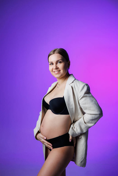 A slim pregnant woman in a long jacket and underwear stands in the studio on a white background, holding her hand on her stomach. Women's underwear for pregnant women - 写真・画像