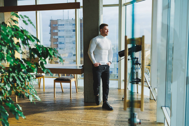 Un giovane bell'uomo in un maglione bianco è in piedi in un ampio ufficio luminoso. Il concetto di una persona moderna di successo. Giovane ragazzo sorridente in open office - Foto, immagini
