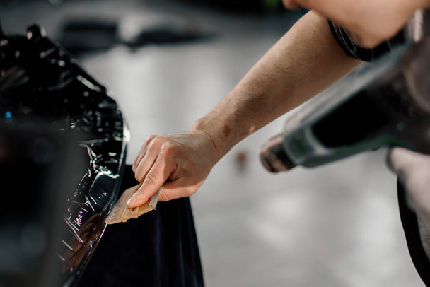 A worker covers a car spoiler with a film in the workshop using hair dryer and spatula close-up Car detailing and care - Photo, Image