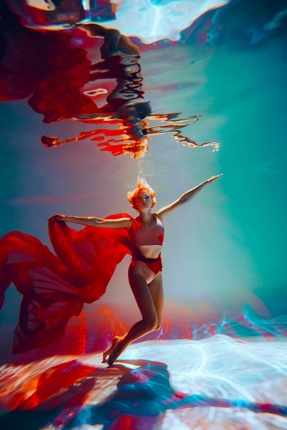 Kunstwerk. Een slank, gebruind meisje met een atletisch figuur en blond haar, met rood materiaal en licht ondergoed, in een ballet poseren onder water in het zwembad. Esthetisch beeld voor uw ontwerp of decoratie. - Foto, afbeelding