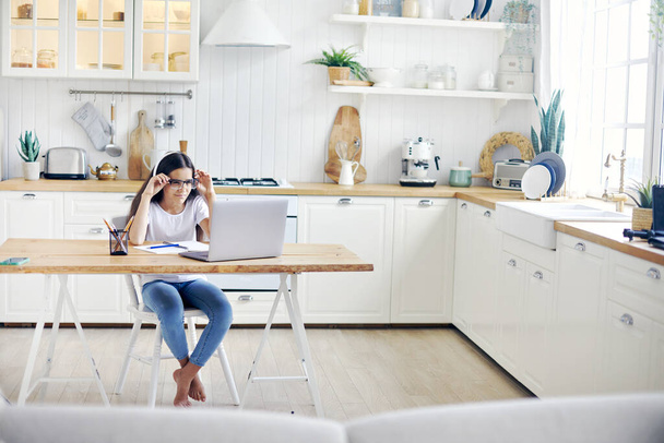 Pretty girl 12s wear headphones and glasses make task, studying online, experiences vision problems, squinting, having eyesight falls, health problems sit at table in kitchen. Poor vision, modern tech - Fotoğraf, Görsel