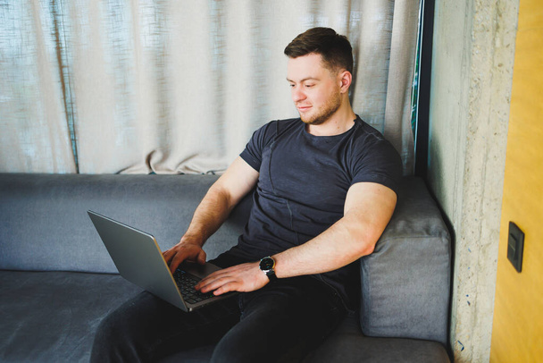 Smiling man in t-shirt sitting on sofa, typing on netbook, working remotely on startup as freelancer, looking at laptop and smiling - Foto, Bild