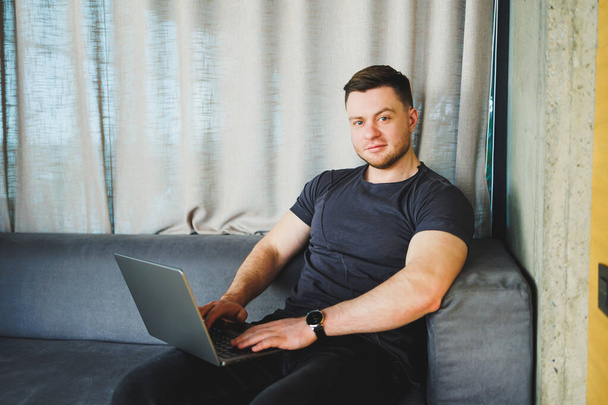 Homem sorridente em camiseta sentado no sofá, digitando no netbook, trabalhando remotamente na inicialização como freelancer, olhando para laptop e sorrindo - Foto, Imagem