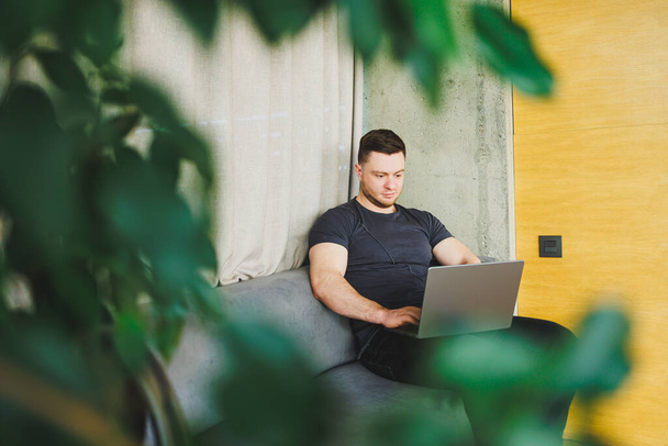 Hymyilevä mies t-paidassa istuu sohvalla, kirjoittaa netbook, työskentelee etänä käynnistyksen freelancerina, katselee kannettavaa tietokonetta ja hymyilee - Valokuva, kuva