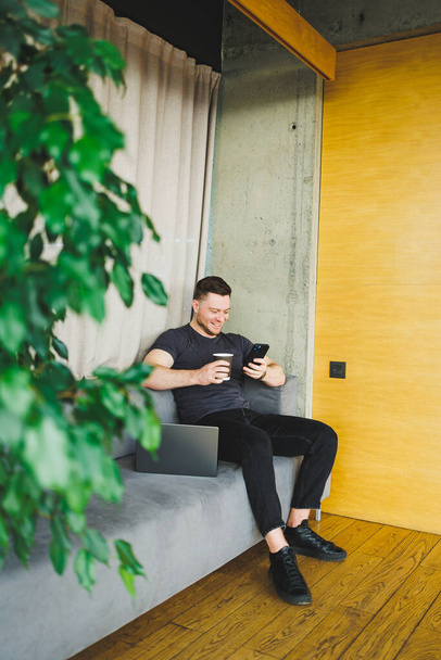 Επιτυχημένος νεαρός άνδρας με t-shirt κάθεται στον καναπέ με τηλέφωνο και πίνοντας καφέ, εργάζεται εξ αποστάσεως κατά τη διάρκεια της εκκίνησης ως ελεύθερος επαγγελματίας, κοιτάζοντας το φορητό υπολογιστή και χαμογελώντας - Φωτογραφία, εικόνα