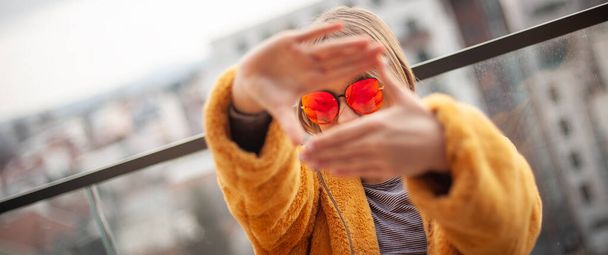 Capture Moments. Young girl in a yellow fur coat and red glasses creates a frame with hands, city in background. - Fotó, kép