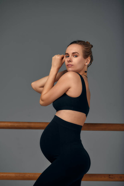Pregnant woman posing near barre in ballet studio in black leggings and a top on a gray background. - Fotoğraf, Görsel
