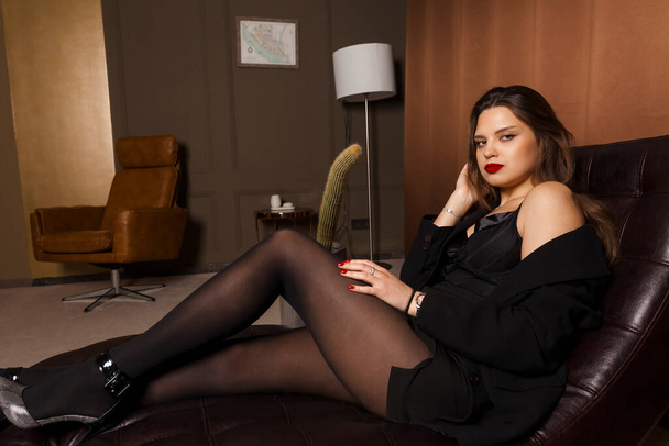 Una joven y atractiva secretaria en medias, vestida formal y sensual, se sienta en una silla de oficina con el pelo suelto, mirando a la cámara. Decoración de la oficina y un cactus alto sobre un fondo. - Foto, imagen