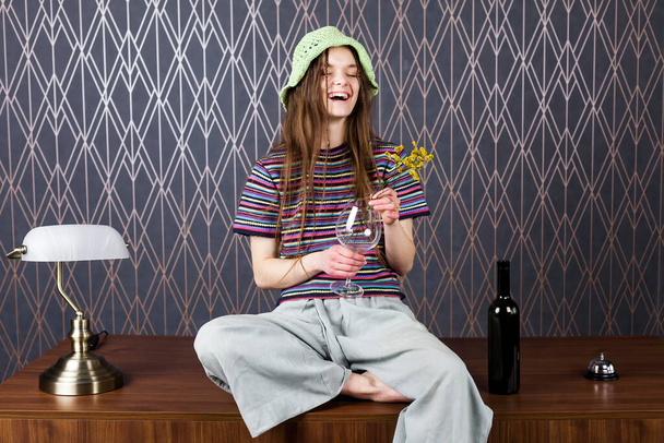 Una graziosa hippie dai capelli lunghi e sorridente vestita con t-shirt a righe multicolore e pantaloni larghi e larghi, seduta su un tavolo con fiori. Concetto di natura e bellezza. - Foto, immagini