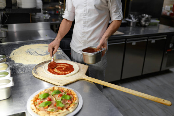 Erkek aşçı pizza tabanına domates ezmesi sürüyor. Lezzetli tariflerle geleneksel İtalyan yemek pişirme süreci - Fotoğraf, Görsel