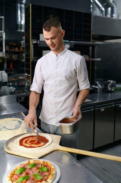 Male chef spreading tomato paste on pizza base. Traditional Italian dish baking process by delicious recipe - Foto, afbeelding