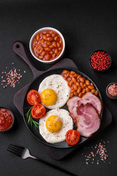 Delicious nutritious English breakfast with fried eggs, tomatoes, bacon and beans on a dark concrete background - Valokuva, kuva