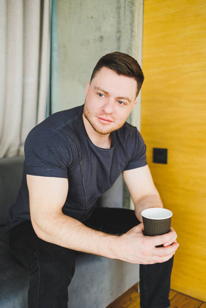 Handsome young man in t-shirt sitting on sofa and drinking coffee, working remotely during startup as a freelancer, looking at laptop and smiling - Fotó, kép