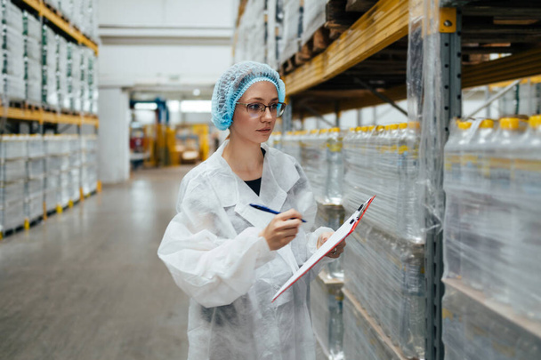 Female worker in protective workwear working in medical supplies research and production factory and checking packed canisters of distilled water before shipment. Inspection quality control. Industrial warehouse. - Foto, immagini
