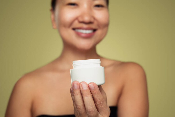Soft focus on white blank jar of cream in hand of smiling Asian model on green background - Foto, Imagem