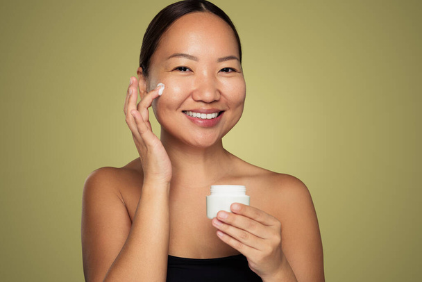 Positive Asian female applying moisturizing cream on cheek while looking at camera against green background - Fotoğraf, Görsel