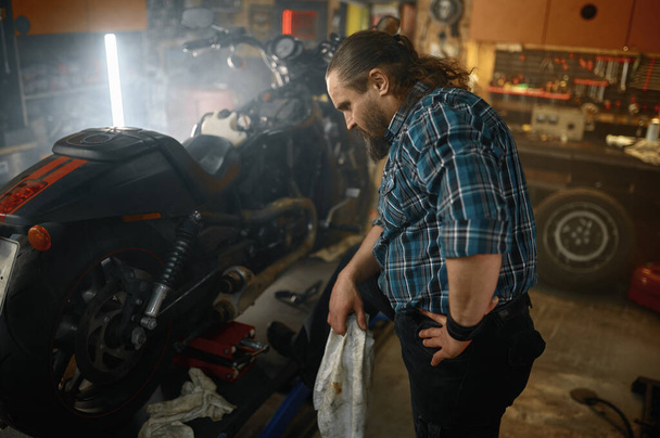 Brutale motorrijder veegt zijn handen af tijdens het repareren van motorfiets motor in de reparatiewerkplaats. Baard mannelijke monteur werken aan motor onderhoud concept - Foto, afbeelding