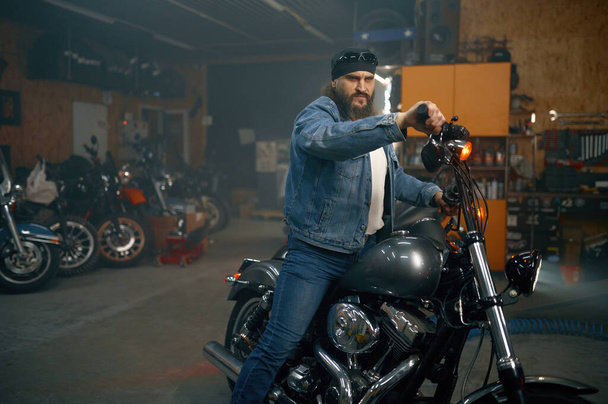 Красивый бородатый мужчина тестирует новый мотоцикл в гаражном магазине. Байкер выбирает новый автомобиль в автомагазине - Фото, изображение
