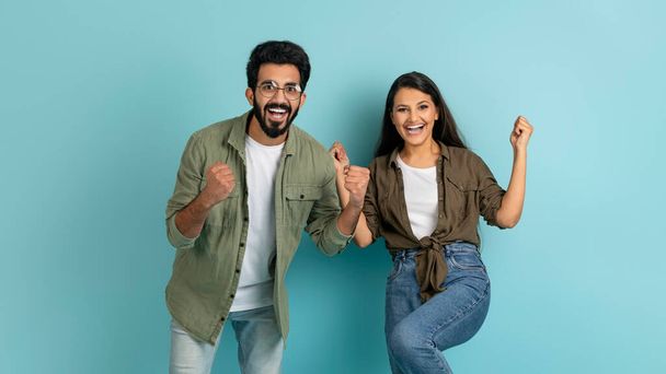 Émotionnel joyeux jeune élégant couple multiethnique homme et femme dans un geste décontracté sur fond de studio bleu, serrant les poings et souriant à la caméra, célébrer le succès ensemble - Photo, image