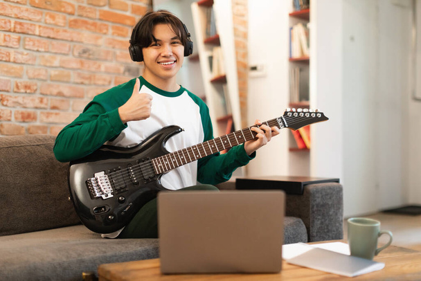 Japonés adolescente chico jugando guitarra eléctrica gestos pulgares hacia arriba sonriendo a la cámara, aprendiendo a jugar en línea en el ordenador portátil sentado en casa, el uso de auriculares. Gadgets musicales. Enfoque selectivo - Foto, Imagen