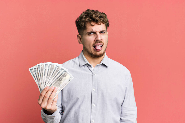 young adult caucasian man feeling disgusted and irritated and tongue out. dollar bank notes concept - Zdjęcie, obraz