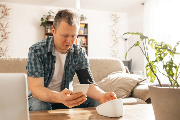 Un hombre mide su presión arterial en casa con un monitor de presión arterial esposado y registra sus resultados en su teléfono. Control de salud en casa. Concepto de hipotensión o hipertensión. - Foto, imagen