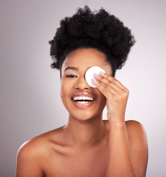 Μαύρη γυναίκα, περιποίηση δέρματος και βαμβάκι στο μάτι στο στούντιο με καθαρισμό, αφαίρεση μακιγιάζ ή γέλιο από το παρασκήνιο. Νεαρό μοντέλο, σκουπίστε και καθαρίστε το δέρμα για φυσική λάμψη, κωμικό χαμόγελο ή καλλυντική υγεία για αυτοπεριποίηση. - Φωτογραφία, εικόνα