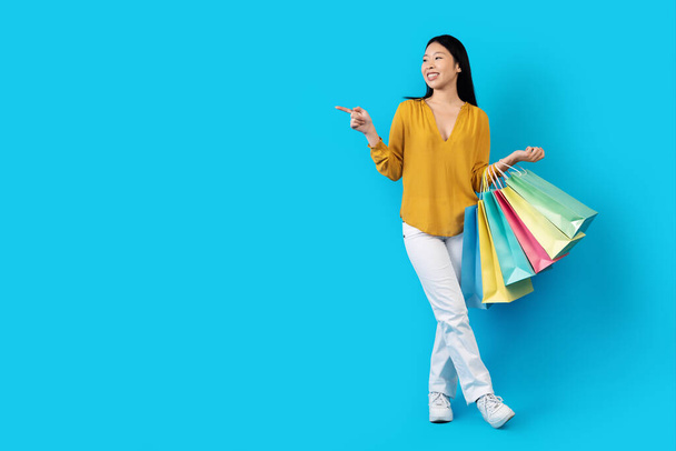 Sonriendo feliz atractiva dama coreana joven de pelo largo en casual shopaholic mostrando espacio libre para la publicidad sobre fondo de estudio azul, hembra llevando bolsas de compras de colores, longitud completa - Foto, imagen