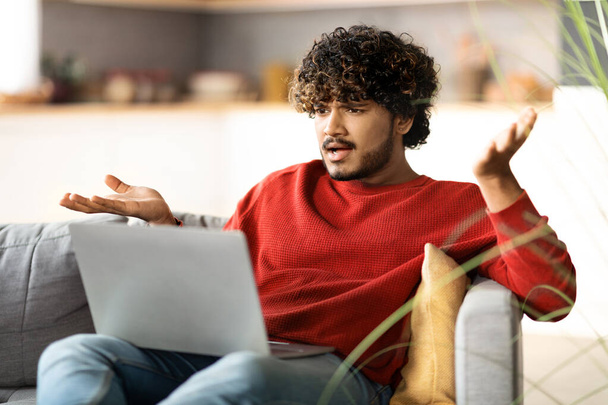 Gestresster junger Inder, der zu Hause Probleme mit dem Laptop hat, ängstlicher Ostler, der auf den Computerbildschirm blickt und emotional gestikuliert, während er im Wohnzimmer auf der Couch sitzt, Nahaufnahme - Foto, Bild