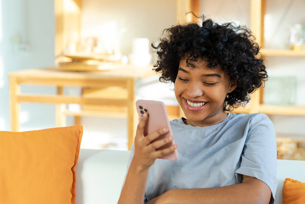 Excited happy young black african american woman holding cell phone laughing feeling joy getting mobile message. Overjoyed girl laughing aloud sitting on couch watching funny video reading news - 写真・画像