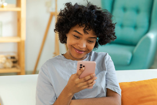 Afrykańska dziewczyna trzyma ekran dotykowy smartfona wpisując przewijanie strony w domu. Kobieta z telefonem komórkowym surfing internet za pomocą aplikacji social media gry. Zakupy online Internet Wiadomości Uzależnienie od telefonów komórkowych - Zdjęcie, obraz