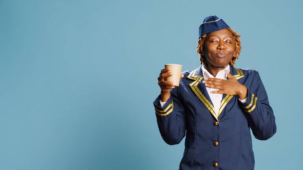 Cheerful stewardess drinking cup of coffee in studio, enjoying caffeine beverage on background. Young aircrew member posing with refreshment or drink on camera, flying airplane. - Photo, Image