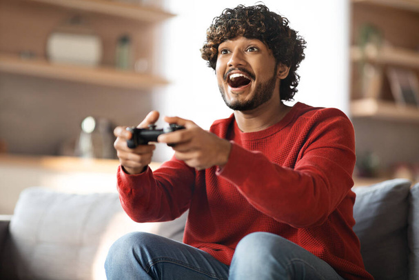 Családi móka. Vidám fiatal indiai srác játszik videojátékok otthon, boldog érzelmi keleti férfi segítségével Joystick és izgalmas, miközben ül a kanapén a nappaliban, közelkép lövés - Fotó, kép
