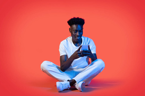 Веселый счастливый красивый молодой черный мужчина в стильной повседневной белой одежде с помощью смартфона, сидя на полу над красным студийным фоном в неоновом свете, скопировать пространство. Зависимость от гаджетов - Фото, изображение