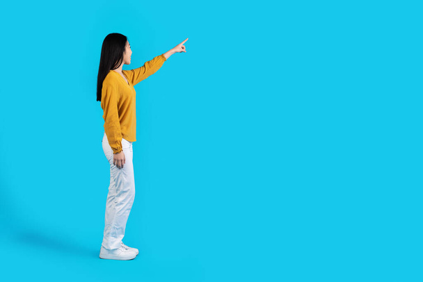 Seitenansicht der schlanken schönen langhaarigen jungen asiatischen Frau in lässigem Outfit, die auf freien Raum für Werbung oder Text zeigt, isoliert auf blauem Studiohintergrund, Aufnahme in voller Länge - Foto, Bild