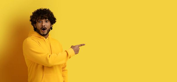 Amazed emotional excited young curly indian guy in yellow hoodie pointing at copy space for shocking advertisement, isolated on yellow studio background, web-banner, panorama - Fotografie, Obrázek