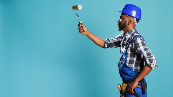 Industrial worker in uniform painting walls with roller, using renovation instrument over blue backdrop. Professional craftsman with hardhat working with rolling paint brush to change color. - 写真・画像