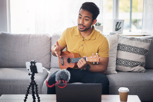 Podcast, guitarra e telefone com um homem online para conversar e treinar durante a aula de streaming ao vivo. ásia masculino pessoa falando no casa sofá com um ukulele como conteúdo criador ensinando música no educação blog. - Foto, Imagem