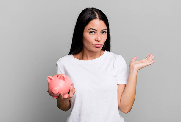 hispanic pretty woman feeling puzzled and confused and doubting with a piggy bank - Foto, Bild