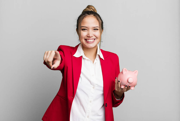 hispanic pretty woman pointing at camera choosing you with a piggy bank - Foto, imagen