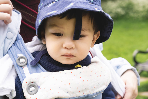 Nahaufnahme eines männlichen Säuglings, der von seinen nicht erkennbaren Eltern auf einer Babytrage getragen wird und einen blauen, deplatzierten Hut trägt. Er ist traurig und blickt nach unten, fühlt sich unwohl. - Foto, Bild