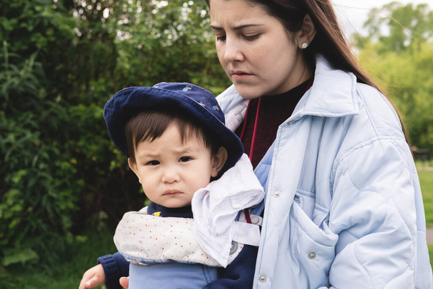 Ein männlicher Säugling blickt in die Kamera, während er von seiner Mutter mit einer Babytrage in blauer Kleidung getragen wird. Ernste und beunruhigende Mimik. Multikulturelle und multirassische Familie. - Foto, Bild