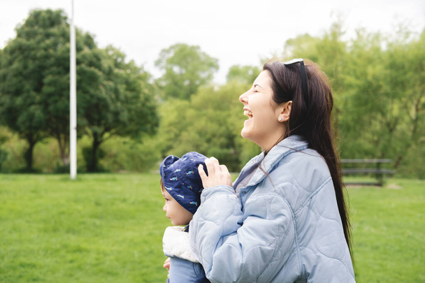 Мати голосно сміється і носить свого чоловічого немовляти з дитячим носієм, гуляючи в парку навесні і носячи блакитний одяг і сонцезахисні окуляри. Біреальна та мультикультурна сім'я
. - Фото, зображення