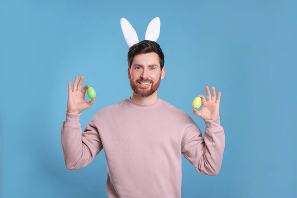 Happy man in cute bunny ears headband holding Easter eggs on light blue background - Fotó, kép