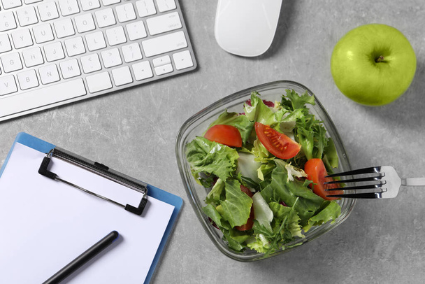Fresh vegetable salad, apple, fork, clipboard and keyboard on grey table at workplace, flat lay. Business lunch - 写真・画像
