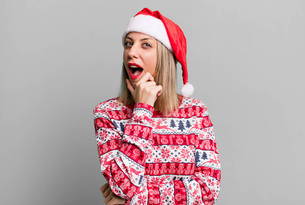 pretty blonde woman with mouth and eyes wide open and hand on chin. christmas and santa hat concept - Foto, imagen