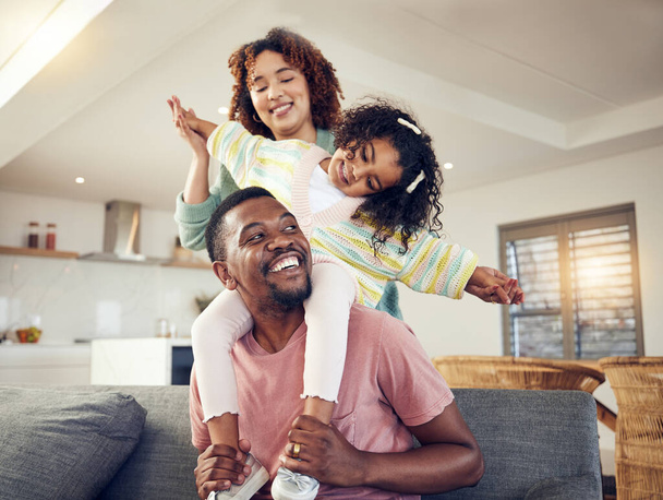 Чорна сім'я, батько і дочка грають з матір'ю на дивані у вітальні для розваги вдома. Щасливий тато, що носить дитину для їзди на поросяті з мамою на кушетці, насолоджуючись відпусткою
. - Фото, зображення