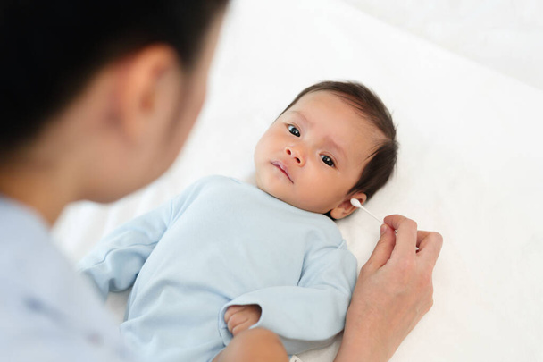 мать, использующая хлопчатобумажный бутон для очистки уха новорожденного ребенка на кровати - Фото, изображение