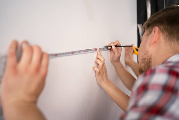 Close-up of a tape measure and pencil in the man's hands. Couple measuring the walls in a room. Repairs in the apartment. Copy space - Photo, Image